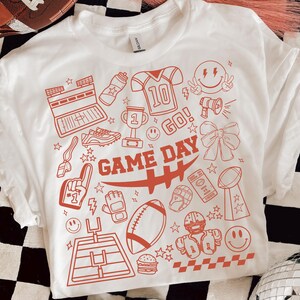 May include: White t-shirt with a red graphic design featuring football-themed illustrations. The design includes a football, helmet, trophy, and the words "GAME DAY". The shirt has a classic crew neck and short sleeves.