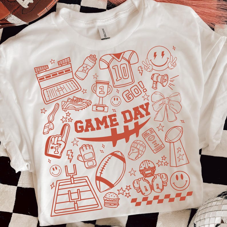 May include: White t-shirt with a red graphic design featuring football-themed illustrations. The design includes a football, helmet, trophy, and the words "GAME DAY". The shirt has a classic crew neck and short sleeves.