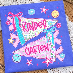 Peut inclure: Un t-shirt bleu avec deux graphismes en forme de fanion portant le texte "KINDER" et "GARTEN" en rose. Le motif comprend des imprim&eacute;s floraux, des rubans roses, des fleurs bleues et roses, des &eacute;toiles jaunes et roses, et deux smileys.