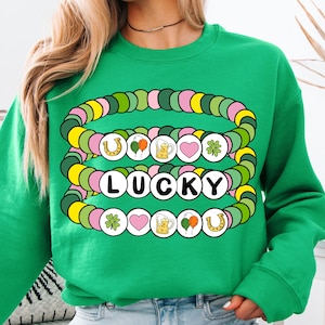 May include: Green sweatshirt with a graphic of layered colorful bracelets with charms, including shamrocks, hearts, beer mugs, horseshoes, and balloons. The word "LUCKY" is spelled out in white letters on the bracelets.