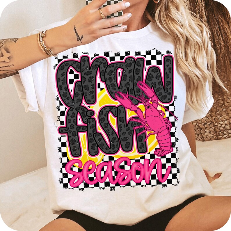 May include: White t-shirt with a black and white checkered background. The text "Crawfish Season" is written in black with a pink crawfish graphic.