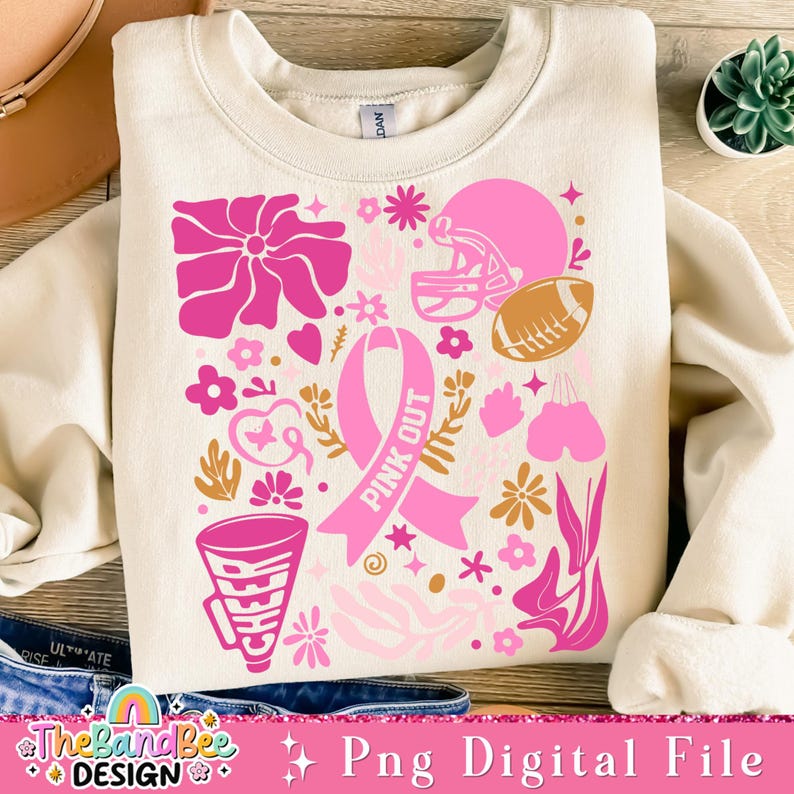 May include: A pink and gold design on a cream colored sweatshirt. The design includes a pink ribbon, flowers, a football, a boxing glove, and a megaphone. The text "Pink Out" is written on the ribbon.