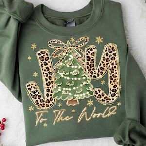 May include: Green sweatshirt with a Christmas tree design. The word "JOY" is in leopard print with gold trim. The Christmas tree is decorated with lights and a bow. The phrase "To The World" is written in gold script.