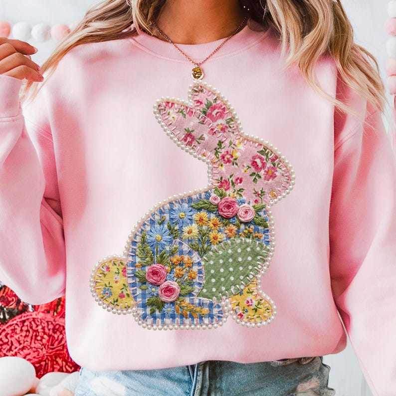 May include: A light pink sweatshirt featuring a patchwork bunny design. The bunny is adorned with floral patterns, including pink roses, blue and yellow flowers, and a polka-dot section. The design is outlined with a pearl-like trim.
