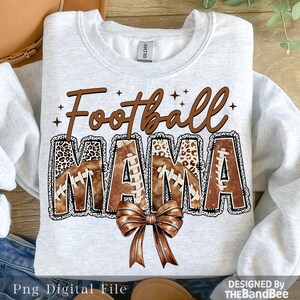 May include: A white sweatshirt with the text "Football Mama" in brown letters with a brown and black football pattern. The letters are outlined in black and have a white glitter effect. A brown bow is below the text.