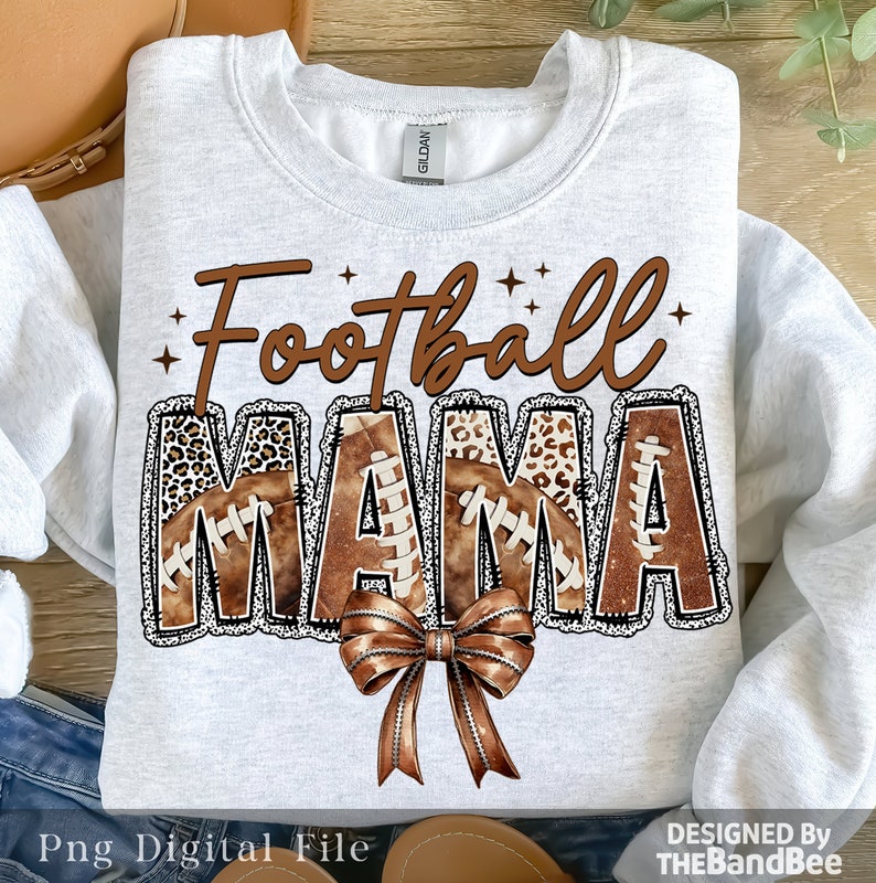 May include: A white sweatshirt with the text "Football Mama" in brown letters with a brown and black football pattern. The letters are outlined in black and have a white glitter effect. A brown bow is below the text.
