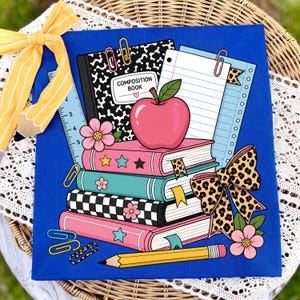 May include: A bright blue fabric square featuring a school-themed graphic with a composition book, a stack of four colorful books, a red apple, a yellow pencil, paper clips, and a large leopard print bow, accented by a yellow striped pencil bow.