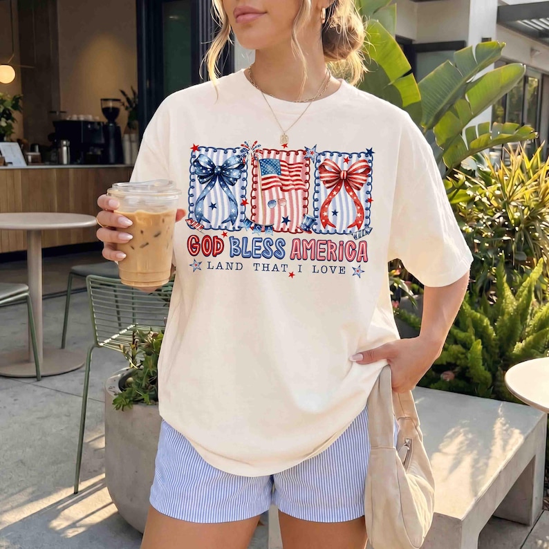 Puede incluir: Camiseta color crema con dise&ntilde;o patri&oacute;tico. El gr&aacute;fico incluye lazos rojos, blancos y azules y una bandera estadounidense, con el texto "GOD BLESS AMERICA LAND THAT I LOVE". La camiseta se combina con pantalones cortos a rayas azules y blancas.