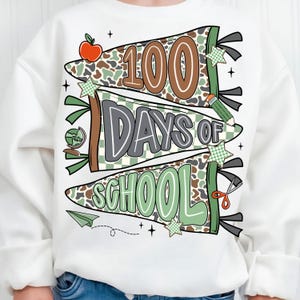 May include: White sweatshirt with a colorful graphic celebrating 100 days of school. The design features pennant banners with the text "100 Days of School" in brown, gray, and green, along with school-themed illustrations like an apple and pencil.