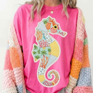 Puede incluir: Una camiseta rosa brillante con un diseño de caballito de mar en patchwork. El caballito de mar está decorado con corales, anclas y patrones de piña. La camiseta se combina con una colorida rebeca a rayas.