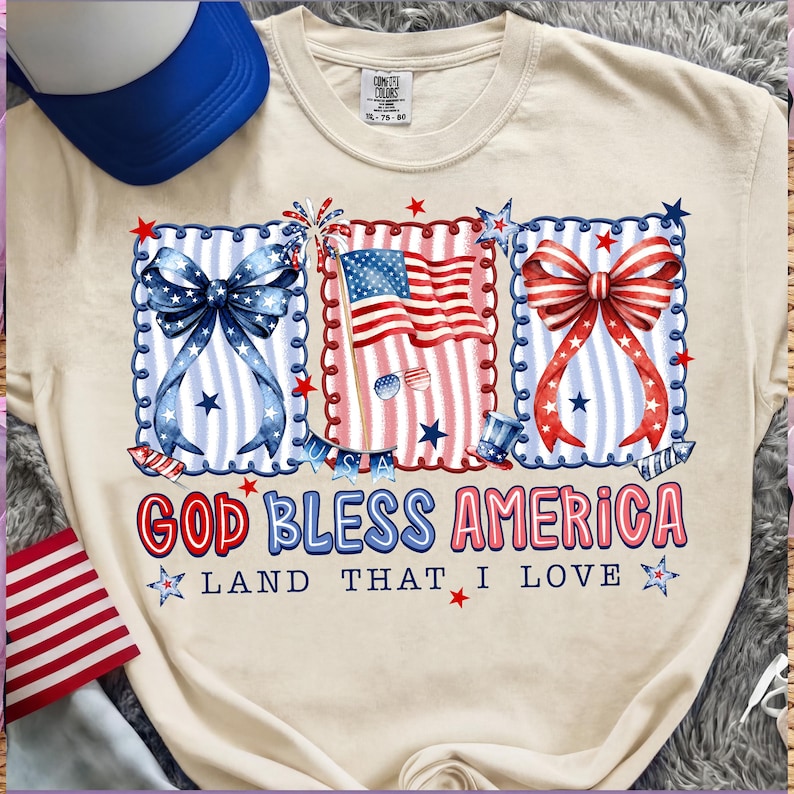 Puede incluir: Camiseta color crema con gr&aacute;ficos patri&oacute;ticos. El dise&ntilde;o incluye lazos azules y rojos, una bandera estadounidense y el texto "GOD BLESS AMERICA LAND THAT I LOVE" en rojo y azul. Una gorra de camionero azul y blanca es visible en la esquina superior izquierda.