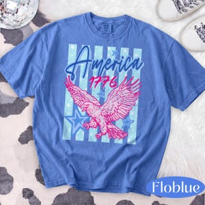 Puede incluir: Camiseta azul con un gr&aacute;fico de &aacute;guila rosa, la palabra "America" y el a&ntilde;o "1776". El dise&ntilde;o incluye rayas y estrellas azules. La camiseta tiene cuello redondo y mangas cortas. El material parece suave y c&oacute;modo.