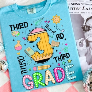 Pu&ograve; includere: T-shirt azzurra con una grafica colorata per la terza elementare. Il design include un grande numero 3 a forma di matita, illustrazioni a tema scolastico e le parole "THIRD GRADE".