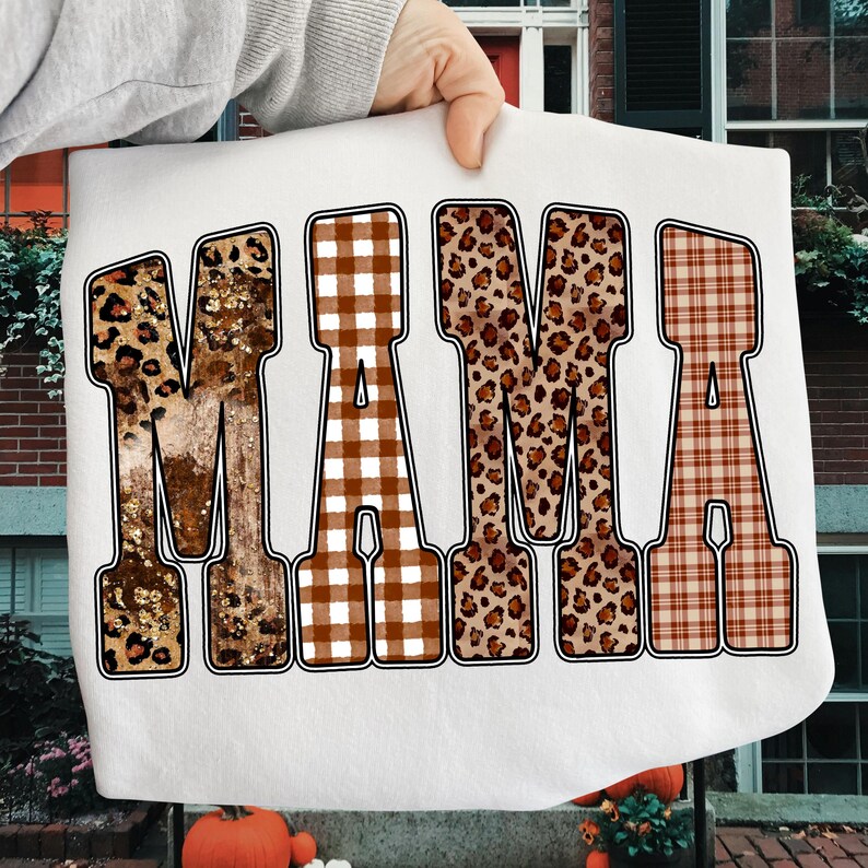 May include: White sweatshirt featuring the word "MAMA" in large, bold letters. Each letter has a unique pattern: leopard print, brown gingham, leopard print, and a plaid design. The letters are outlined in black.