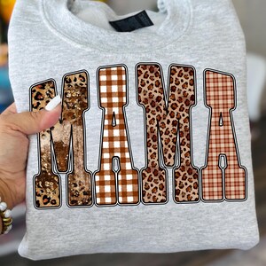 May include: A light gray sweatshirt with the word "MAMA" in large, bold letters. The letters have a mix of animal print, plaid, and glitter patterns. The sweatshirt is folded.