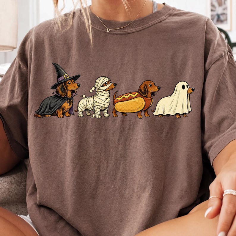 Peut inclure: Un t-shirt marron &agrave; manches courtes avec un motif repr&eacute;sentant quatre teckels en costumes d'Halloween. Les chiens sont d&eacute;guis&eacute;s en sorci&egrave;re avec une cape et un chapeau noirs, en momie avec des bandages blancs, en hot-dog et en fant&ocirc;me sous un drap.