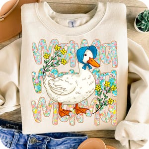 May include: Cream-coloured sweatshirt with a cartoon goose wearing a blue bonnet and floral accents. The word "MAMA" is printed in a floral pattern. The goose has an orange beak and feet. The sweatshirt is a casual style.