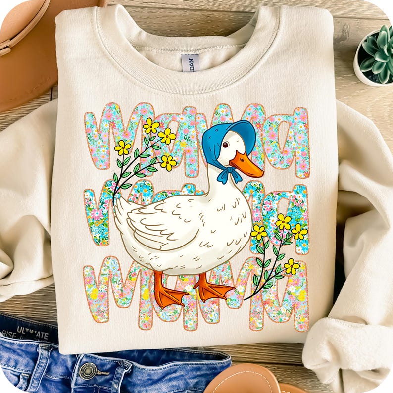 May include: Cream-coloured sweatshirt with a cartoon goose wearing a blue bonnet and floral accents. The word "MAMA" is printed in a floral pattern. The goose has an orange beak and feet. The sweatshirt is a casual style.