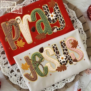 May include: Two embroidered shirts, one red and one white, with the words "PRAY" and "JESUS". The letters are decorated with fall-themed elements like leaves, acorns, and leopard print. The top left corner of the red shirt has the text "ORIGINAL DESIGN".