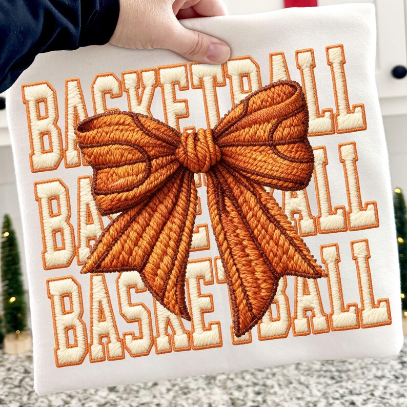 May include: White sweatshirt with the word "BASKETBALL" embroidered in orange block letters. A large, detailed orange bow is embroidered in the center. The bow has a textured appearance, with darker brown outlining. The sweatshirt is held by a hand.