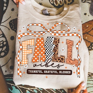 May include: A beige sweatshirt with the words "FALL VIBES" in orange, black, and white lettering. The word "FALL" has a gingham pattern, a lightning bolt, and a leopard print. The text "THANKFUL, GRATEFUL, BLESSED" is below.