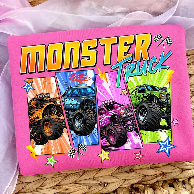 May include: Pink t-shirt featuring the words "MONSTER TRUCK" in bold, outlined letters. The design includes four colorful monster trucks with flames, lightning bolts, and stars. The background has a comic book style with vibrant colors.