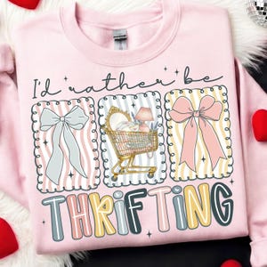 以下が含まれることがあります： 「I'd rather be thrifting」の文字が入ったピンク色のスウェットシャツ。リボン、商品でいっぱいのショッピングカート、カラフルな文字で「thrifting」という言葉のイラストがデザインされています。スウェットシャツはクルーネックです。