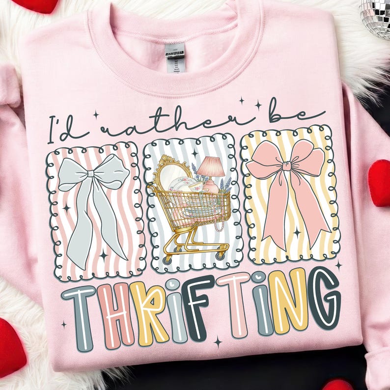 May include: Pink sweatshirt with the phrase "I'd rather be thrifting." The design includes illustrations of bows, a shopping cart filled with items, and the word "thrifting" in colorful letters. The sweatshirt has a crew neck.