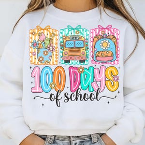 K&ouml;nnte beinhalten: Wei&szlig;es Sweatshirt mit einem farbenfrohen Grafikdesign, das "100 Schultage" feiert. Das Design zeigt Illustrationen von Schulmaterialien, einem Schulbus und einem Rucksack, alle mit floralen Akzenten.