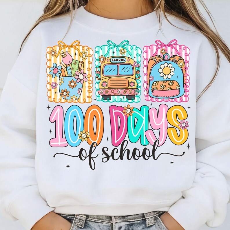 K&ouml;nnte beinhalten: Wei&szlig;es Sweatshirt mit einem farbenfrohen Grafikdesign, das "100 Schultage" feiert. Das Design zeigt Illustrationen von Schulmaterialien, einem Schulbus und einem Rucksack, alle mit floralen Akzenten.