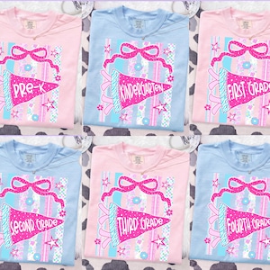 May include: Six pastel-colored t-shirts with school grade graphics. Each shirt features a pennant design with text indicating the grade level, Pre-K through Fourth Grade. The pennants are pink with blue accents and star details.