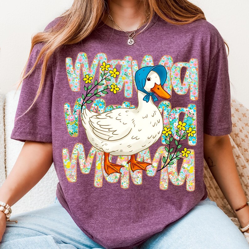 May include: A purple t-shirt with the word "Mama" in a floral design. A white duck wearing a blue bonnet is in the centre, surrounded by a floral wreath. The duck has an orange beak and feet. The shirt is a casual style.