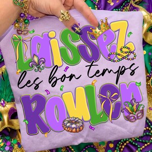 May include: A light purple t-shirt with the phrase "Laissez les bon temps rouler" in green, yellow, and purple. The design includes a crown, beads, a fleur-de-lis, a mask, and a king cake, all in festive colors.
