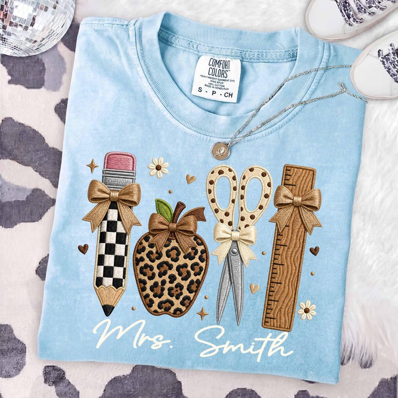 May include: A light blue t-shirt featuring an embroidered design of a checkered pencil, a leopard print apple, scissors with a polka dot pattern, and a wooden ruler, each accented with a tan bow. The text Mrs. Smith is embroidered in white.