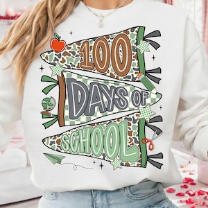 May include: White sweatshirt with a colorful graphic celebrating 100 days of school. The design features banners with the words "100 Days of School" in brown, gray, and green, along with school-themed illustrations like an apple, pencil, and globe.