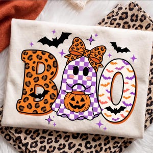 Puede incluir: Una camiseta color crema con un gráfico de la palabra BOO. La letra B tiene un estampado de leopardo naranja, la O es un fantasma con un patrón de cuadros morados y un lazo de leopardo naranja, y la última O tiene un fondo blanco con murciélagos.