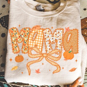 May include: A beige sweatshirt with the word "MAMA" in orange, floral, and gingham patterns. A large orange bow is below the word, with pumpkins and leaves. The design has a fall theme.