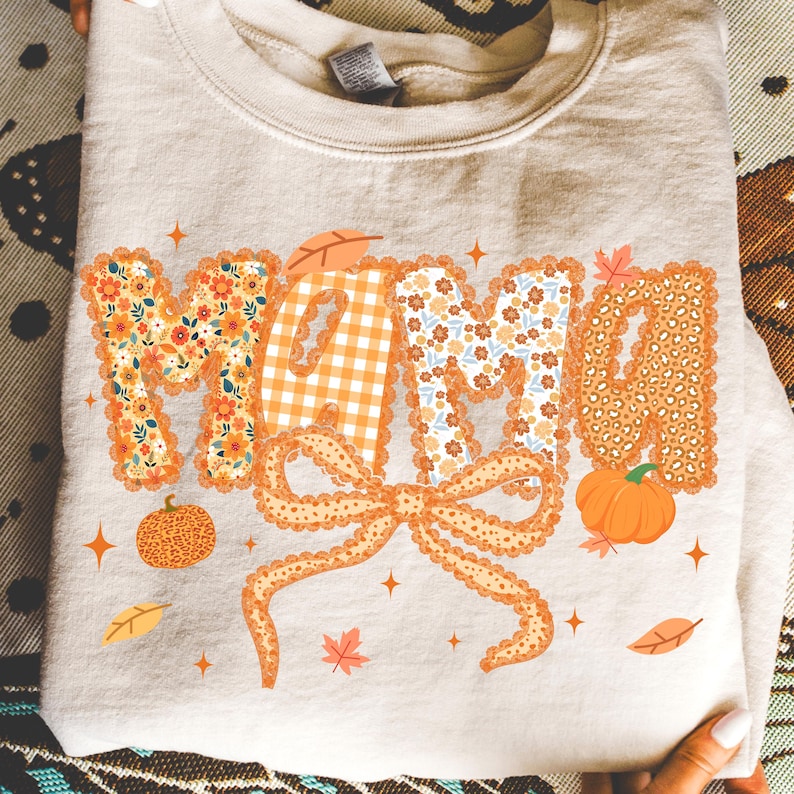 May include: A beige sweatshirt with the word "MAMA" in orange, floral, and gingham patterns. A large orange bow is below the word, with pumpkins and leaves. The design has a fall theme.
