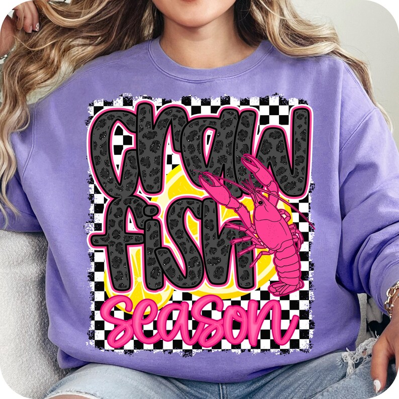 May include: Purple sweatshirt with a black and white checkered background. The text "Crawfish Season" is written in black with a leopard print pattern. A pink crawfish is in the center of the design.