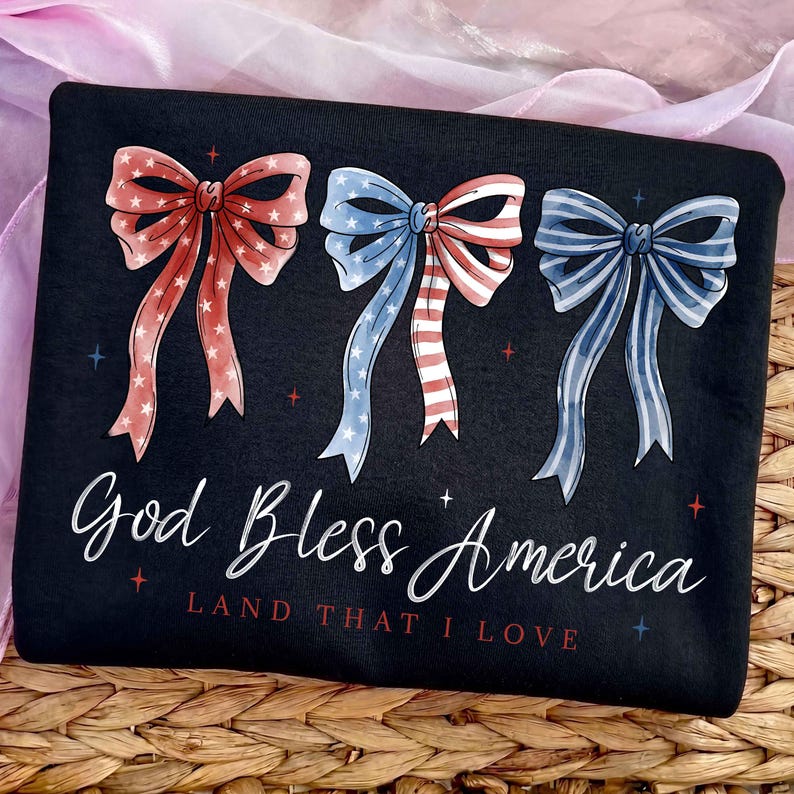 May include: Black t-shirt featuring three patriotic bow designs in red, white, and blue. The text "God Bless America" and "Land That I Love" are written in white script. The bows have star and stripe patterns.