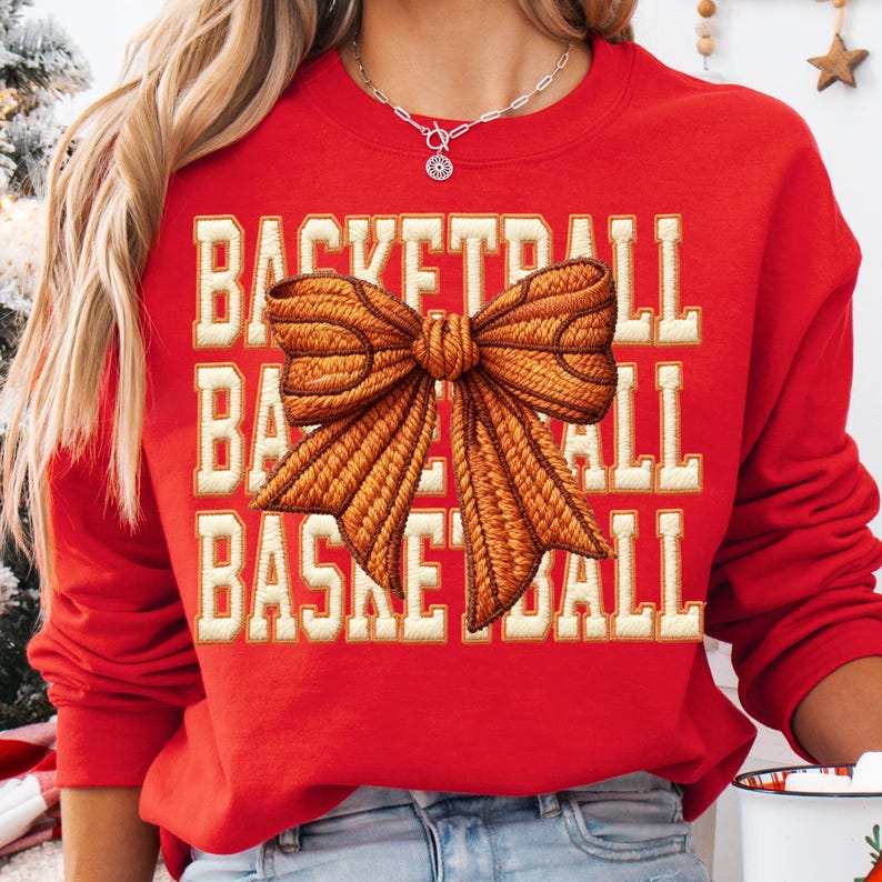May include: A red sweatshirt with the word "BASKETBALL" in white block letters. A large, detailed bow in shades of brown is centered on the sweatshirt. The sweatshirt is a casual, comfortable style.