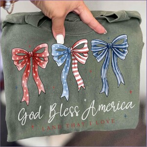 May include: Olive green sweatshirt featuring three patriotic bow designs in red, white, and blue. The bows are watercolor-style with star and stripe patterns. The text "God Bless America, Land That I Love" is printed below the bows.