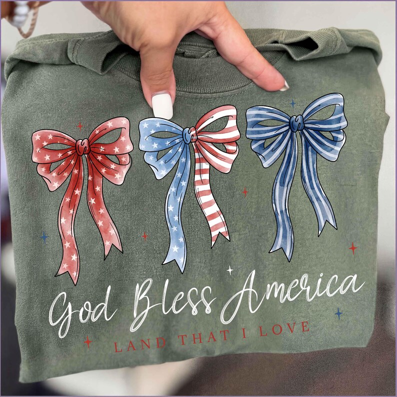 May include: Olive green sweatshirt featuring three patriotic bow designs in red, white, and blue. The bows are watercolor-style with star and stripe patterns. The text "God Bless America, Land That I Love" is printed below the bows.