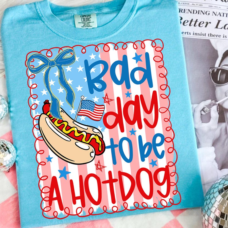 May include: Light blue t-shirt with a graphic that says "Bad day to be a hotdog." The graphic features a cartoon hotdog with an American flag and a blue bow. The background has red and white stripes and blue stars.