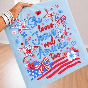 Könnte beinhalten: Hellblaue Decke mit patriotischem Design. Mit roten, weißen und blauen Sternen, Schleifen und floralen Akzenten. Der Text lautet "She loves Jesus and America too". Ideal für Heimdekoration oder als Geschenk.
