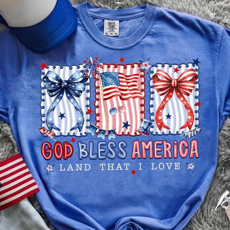 Puede incluir: Una camiseta azul con un dise&ntilde;o patri&oacute;tico. El dise&ntilde;o incluye un lazo azul, una bandera estadounidense y un lazo rojo, todo enmarcado con bordes decorativos y estrellas. El texto dice "GOD BLESS AMERICA" y "LAND THAT I LOVE".