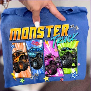 May include: Blue sweatshirt featuring the words "MONSTER TRUCK" in yellow and green. The design includes four colorful monster truck illustrations with flames, lightning bolts, and stars. The trucks are in orange, blue, pink, and green.