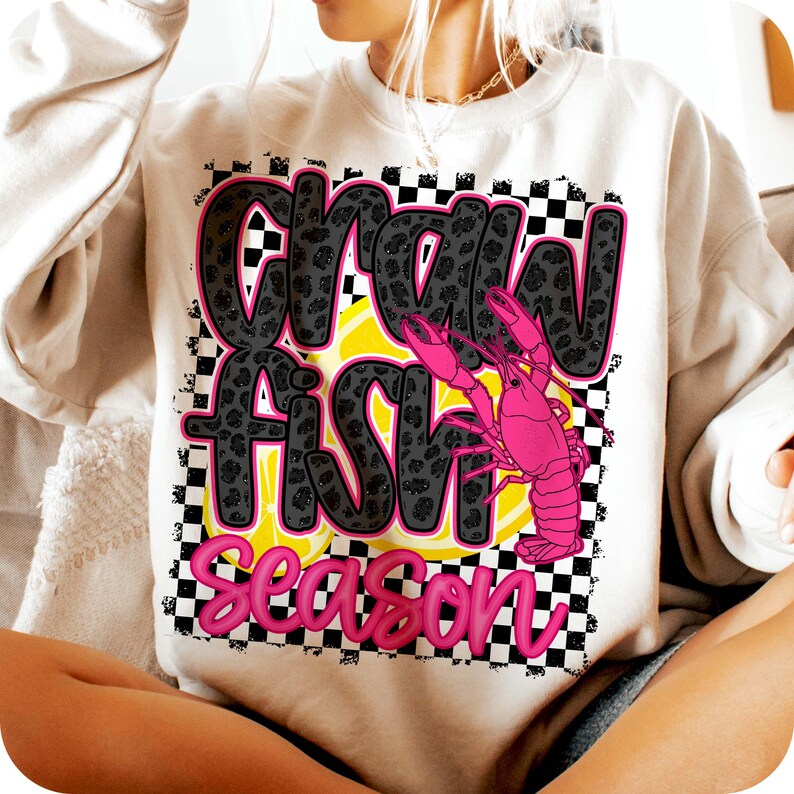 May include: A white sweatshirt with a black and white checkered background. The text "Crawfish Season" is written in pink and black letters with a pink crawfish illustration.