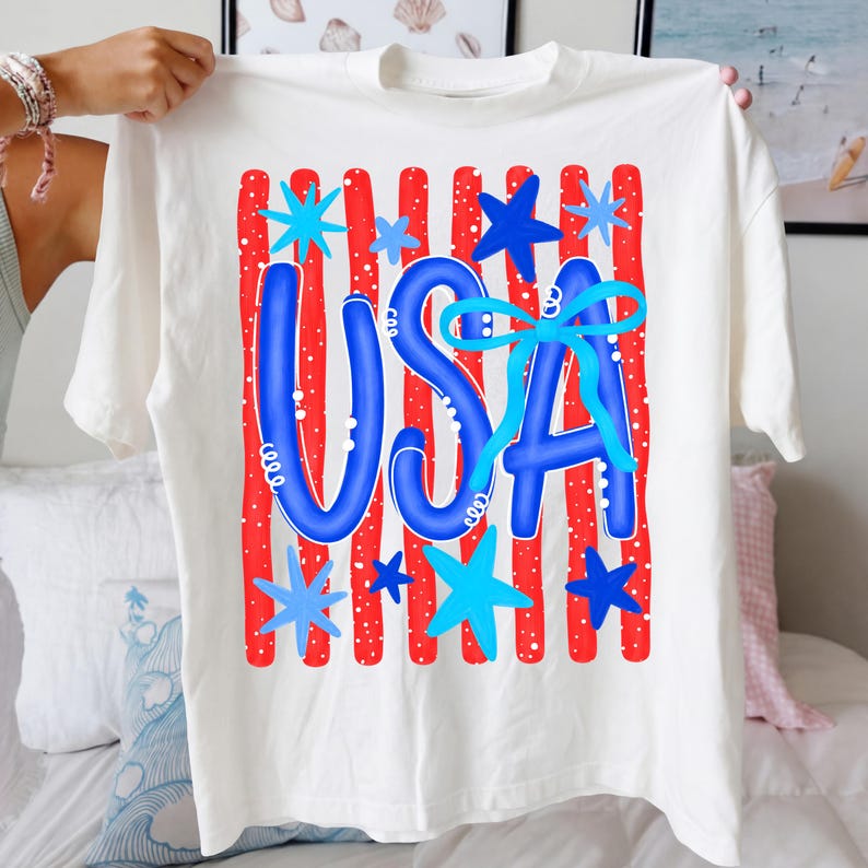 May include: White t-shirt with a patriotic design. The word "USA" is in blue, with a light blue bow and surrounded by blue stars and red stripes. The shirt is held up by a person.