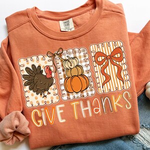 May include: Orange sweatshirt with a Thanksgiving-themed graphic. The graphic includes a turkey, stacked pumpkins, and a bow, all within decorative frames. The words "GIVE THANKS" are printed in a colorful font.